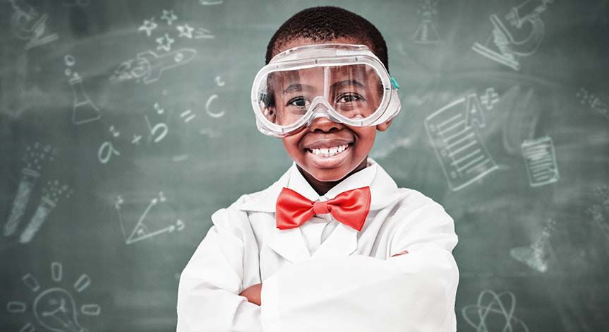 Boy wearing lab coat, red bow tie and safety goggles in a classroom.  Blackboard in the background with different science ico