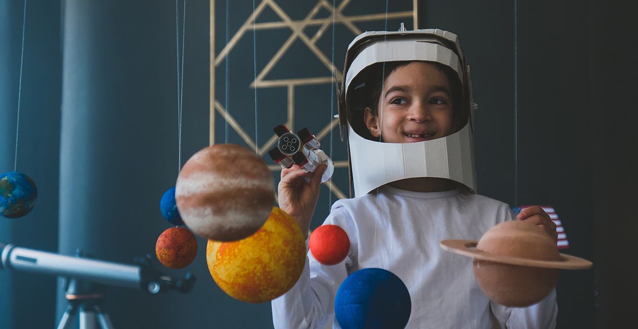 boy wearing cardboard astronaut helmet flying toy rocket through planets, cardboard spaceship rocket in the background