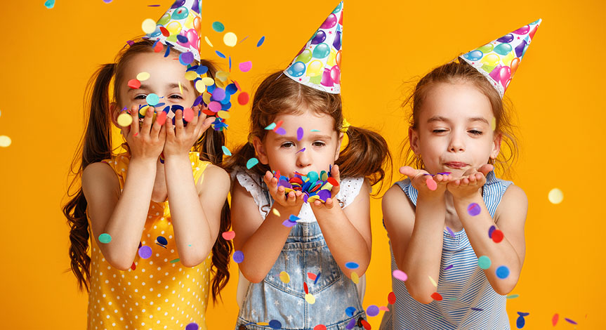 3 girls wearing birthday hats celebrating a birthday with confetti on yellow background