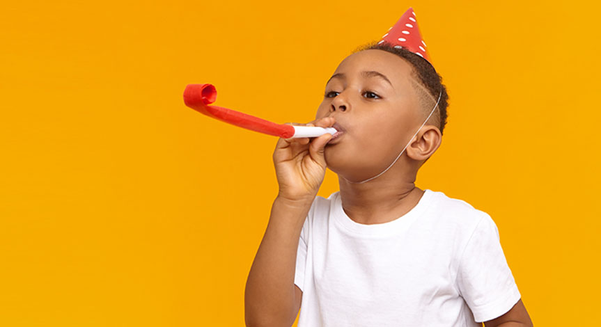 Happy boy celebrating a birthday, posing isolated at blank yellow wall, blowing whistle, Birthday hat on his head